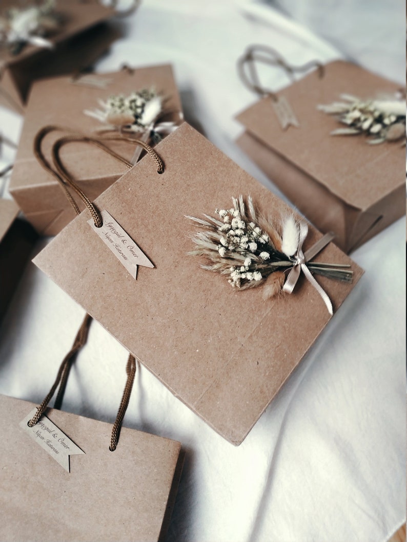May include: Brown paper gift bags with white dried flowers and a brown ribbon. T