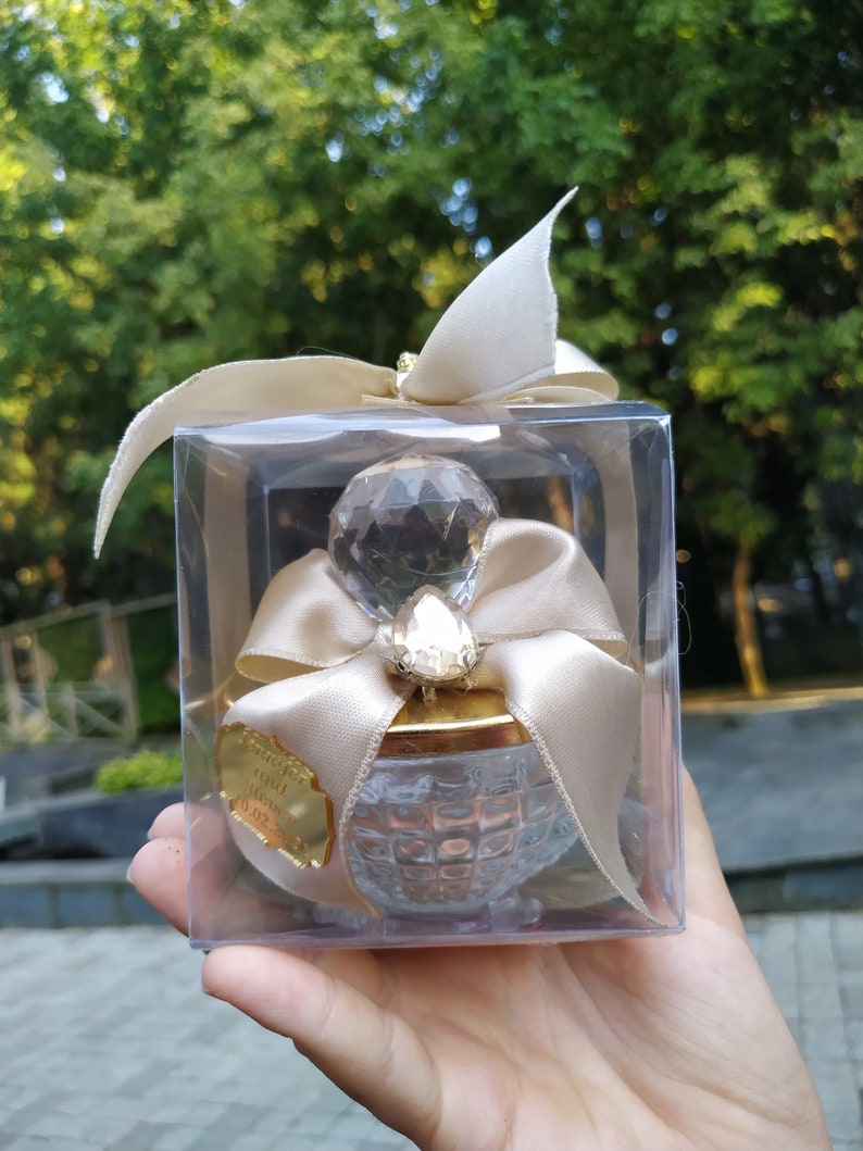May include: A clear glass jar with a gold lid and a crystal-like top. The jar is decorated with a gold ribbon and a small crystal. The jar is in a clear plastic box with a white ribbon.
