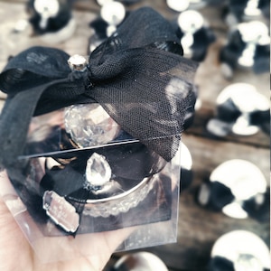 May include: A clear glass box with a black bow and a crystal-like ornament inside. The box is surrounded by other similar boxes with black bows.
