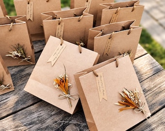 Könnte beinhalten: Eine Ansammlung von braunen Geschenktüten aus Kraftpapier, die jeweils mit einem kleinen Strauß aus Trockenblumen und Weizenähren auf der Vorderseite verziert sind. Jede Tüte hat ein rechteckiges Papieretikett, das mit einer Schnur am Griff befestigt ist.