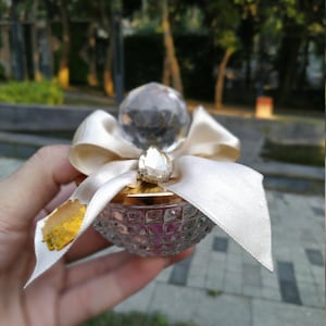 May include: A clear glass bottle with a gold lid and a white ribbon. The bottle is decorated with a crystal-like stopper and has a textured surface.