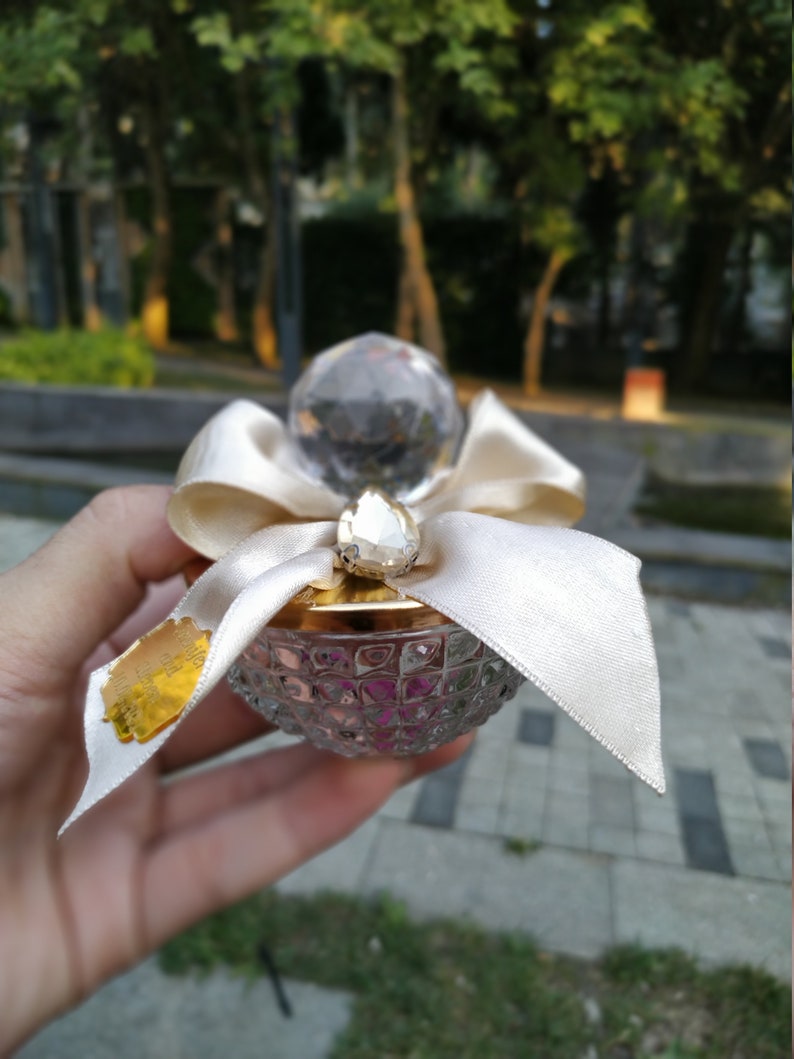 May include: A clear glass bottle with a gold lid and a white ribbon. The bottle is decorated with a crystal-like stopper and has a textured surface.