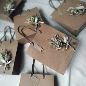 May include: Five brown paper gift bags with white string handles. Each bag has a small bouquet of dried white flowers and greenery attached to the front. The bags have a small tag attached with the text 'Engaged & Cheers'