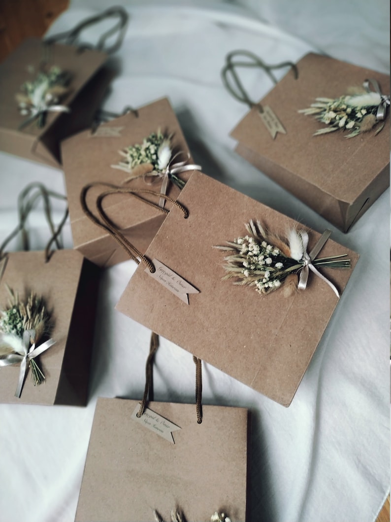 May include: Five brown paper gift bags with white string handles. Each bag has a small bouquet of dried white flowers and greenery attached to the front. The bags have a small tag attached with the text 'Engaged & Cheers'