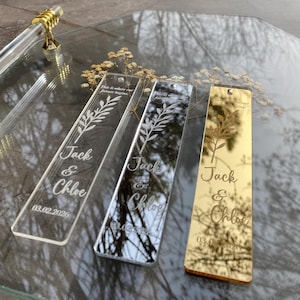 May include: Three rectangular acrylic bookmarks with engraved text and a floral design. The bookmarks are clear, mirrored, and gold. The text reads "Jack & Chloe" and "03.02.2026". The top of each bookmark has the text "This is where our forever begins."