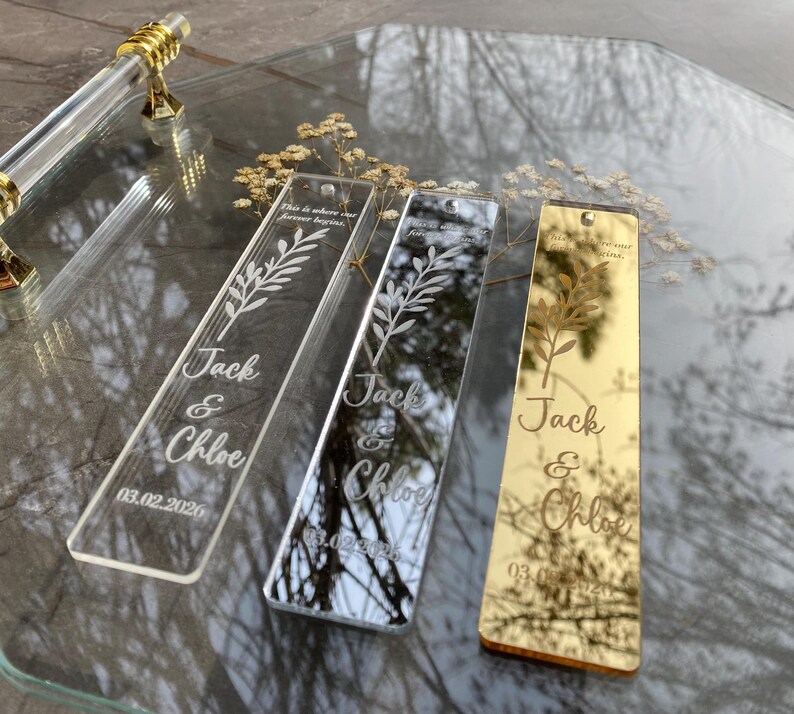 May include: Three rectangular acrylic bookmarks with engraved text and a floral design. The bookmarks are clear, mirrored, and gold. The text reads "Jack & Chloe" and "03.02.2026". The top of each bookmark has the text "This is where our forever begins."