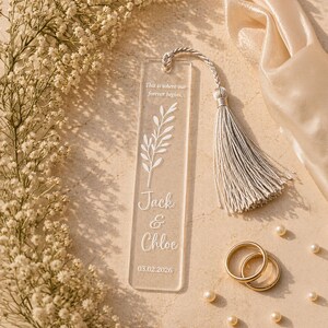 May include: A clear acrylic bookmark with a white floral design and the names "Jack & Chloe" and the date 03.02.2026. The bookmark has a silver tassel and is surrounded by flowers, wedding rings, and pearls.