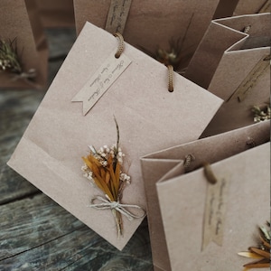 May include: A collection of small brown paper gift bags with twine handles. Each bag has a small tag with the text "Thank You & Cheers" and a small bouquet of dried flowers.