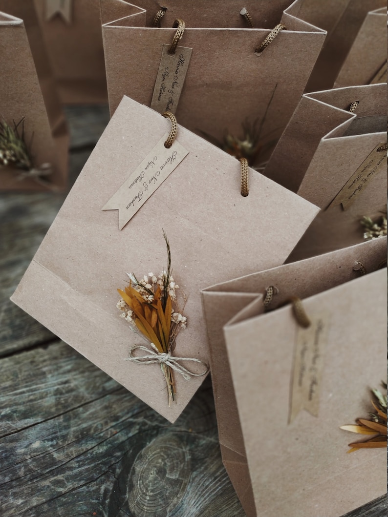 May include: A collection of small brown paper gift bags with twine handles. Each bag has a small tag with the text "Thank You & Cheers" and a small bouquet of dried flowers.