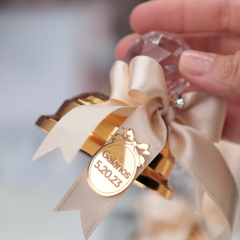 May include: A gold-colored glass jar with a crystal-shaped lid and a beige satin ribbon tied in a bow. The jar has a gold-colored label with the text "Galanos 520 23".