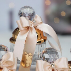 May include: A set of clear glass bottles with gold lids and crystal stoppers. Each bottle is tied with a cream-colored satin ribbon and bow. The bottles have a personalized label with the text "& T 2023".
