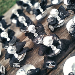 May include: Silver favor boxes with black ribbon and a large clear crystal on top. Each box has a small black tag with white text that reads "HOSNUTE & M. ALI Nikah Meresimi".