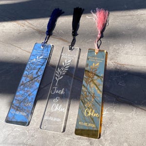 May include: Three personalized acrylic bookmarks with tassels. One is blue, one is clear, and one is gold. Each bookmark has the names "Jack & Chloe" and the date "03.02.2026" engraved. The text "This is where our forever begins" is also included.