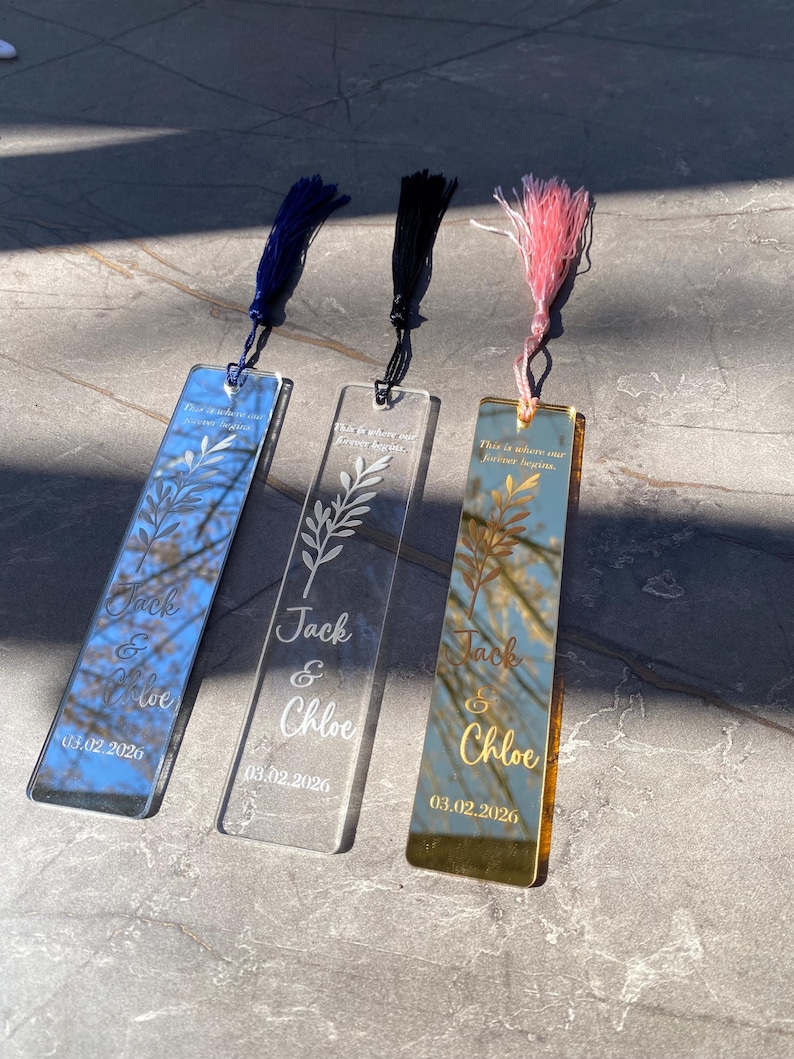 May include: Three personalized acrylic bookmarks with tassels. One is blue, one is clear, and one is gold. Each bookmark has the names "Jack & Chloe" and the date "03.02.2026" engraved. The text "This is where our forever begins" is also included.