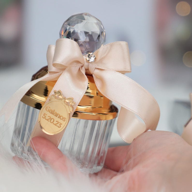 May include: A clear glass jar with a gold lid and a crystal-like top. The jar is decorated with a beige satin ribbon tied in a bow. A gold tag with the text "Galavos 5.20.23" is attached to the jar.