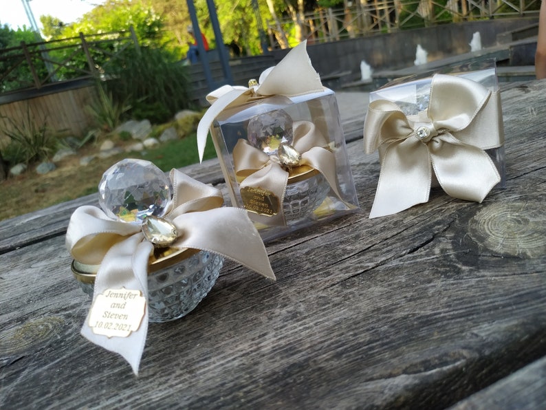 May include: Three small, clear glass containers with gold accents and a crystal-like top. Each container is decorated with a white satin ribbon and bow. One container has a tag with the text "Jennifer and Steven 10.02.2021".