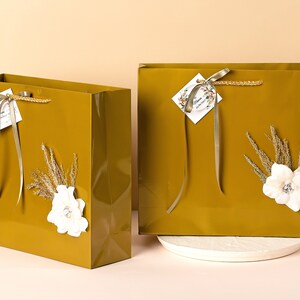 Puede incluir: Dos bolsas de regalo doradas con flores blancas y espigas de hierba seca. Las bolsas tienen una cinta dorada y una peque&ntilde;a etiqueta con la palabra "Happy" en ella.