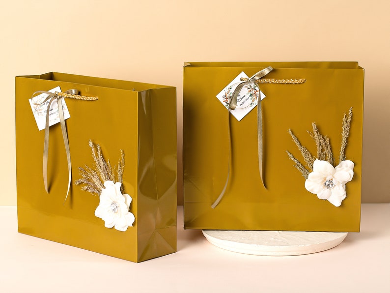 Puede incluir: Dos bolsas de regalo doradas con flores blancas y espigas de hierba seca. Las bolsas tienen una cinta dorada y una peque&ntilde;a etiqueta con la palabra "Happy" en ella.