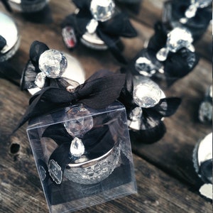 May include: A small, clear glass jar with a silver lid and a large crystal on top. The jar is wrapped in black ribbon and placed in a clear plastic box. The jar is surrounded by other similar jars, all with black ribbon and crystals.