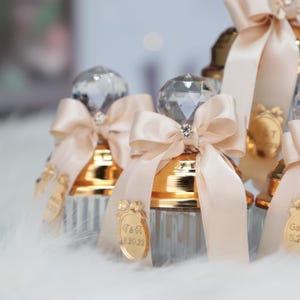 May include: Set of small glass bottles with gold lids and crystal stoppers. Each bottle is tied with a beige satin ribbon and bow. The bottles have personalized labels with the text "T & A 8.20.28" and "G & Z 5.28".