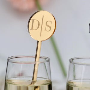 May include: Gold acrylic drink stirrer with a round, mirrored top featuring the initials "D/S" engraved in a sans-serif font.