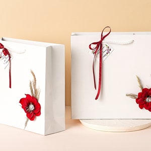 Puede incluir: Dos bolsas de regalo blancas con lazos de cinta roja y adornos de flores rojas. Las bolsas tienen una etiqueta blanca con el texto "Feliz cumplea&ntilde;os" en ellas.