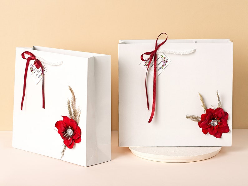 Puede incluir: Dos bolsas de regalo blancas con lazos de cinta roja y adornos de flores rojas. Las bolsas tienen una etiqueta blanca con el texto "Feliz cumplea&ntilde;os" en ellas.