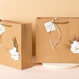 Puede incluir: Dos bolsas de regalo de papel marr&oacute;n con asas de cinta blanca y adornos florales blancos. Una bolsa tiene una peque&ntilde;a etiqueta blanca con el texto "Dearest & Best" en ella.