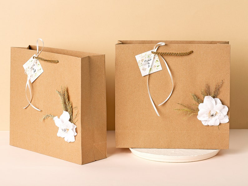 Puede incluir: Dos bolsas de regalo de papel marr&oacute;n con asas de cinta blanca y adornos florales blancos. Una bolsa tiene una peque&ntilde;a etiqueta blanca con el texto "Dearest & Best" en ella.