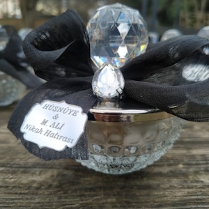 May include: A small, clear glass jar with a silver lid and a crystal-shaped top. The jar is decorated with a black ribbon and a silver tag that reads "H&Uuml;SN&Uuml;YE & M. ALİ Nikah Hatırası".