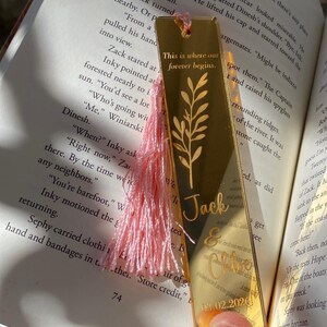 May include: Gold-colored metal bookmark with a pink tassel, personalized with a name and date. The bookmark features an engraved floral design and the text "This is where our forever begins."