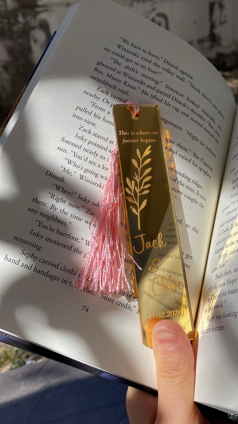 May include: Gold-colored metal bookmark with a pink tassel, personalized with a name and date. The bookmark features an engraved floral design and the text "This is where our forever begins."