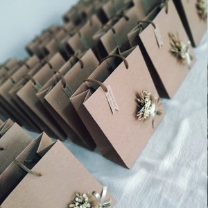 May include: A collection of brown paper gift bags with white string handles. Each bag has a small dried flower arrangement attached to the front. The bags are arranged in rows on a white surface. The text on the tag reads "Jared & Clair, June 2023".