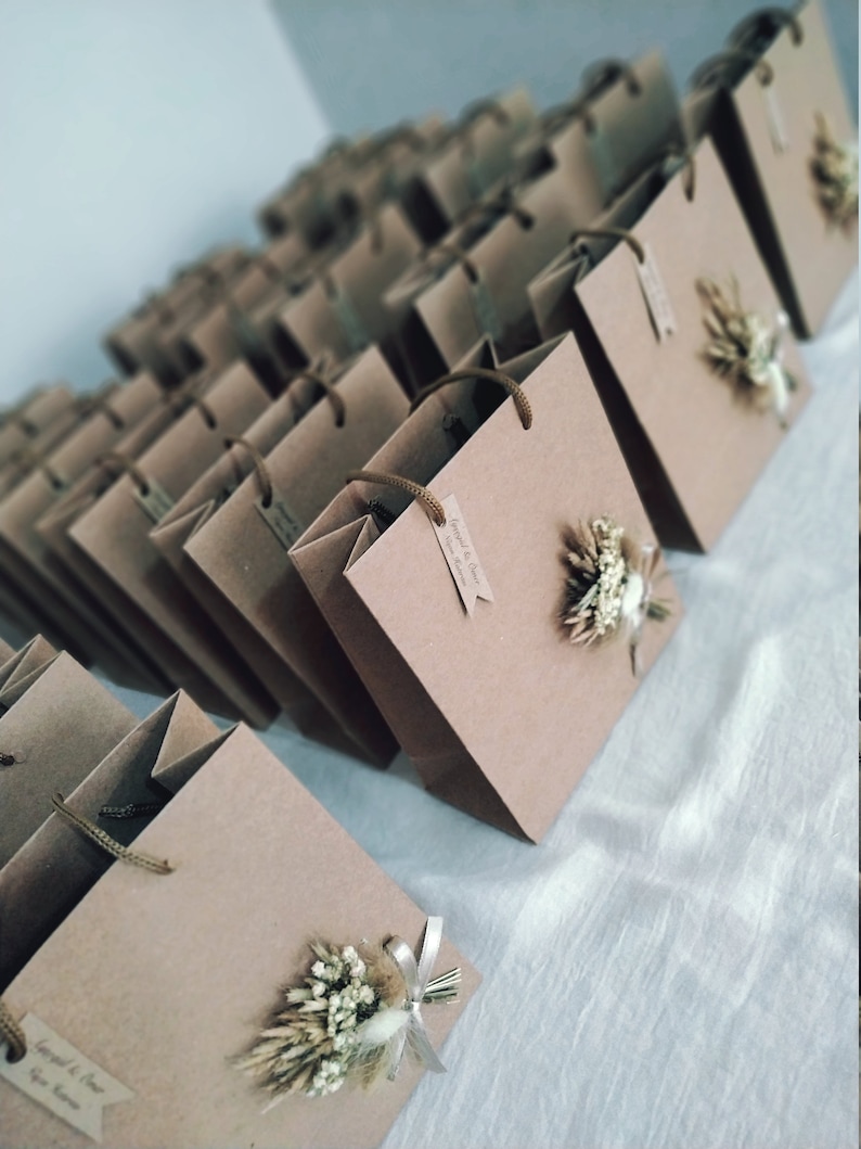 May include: A collection of brown paper gift bags with white string handles. Each bag has a small dried flower arrangement attached to the front. The bags are arranged in rows on a white surface. The text on the tag reads "Jared & Clair, June 2023".