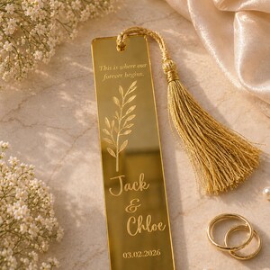 May include: A gold-colored bookmark with a tassel, featuring the engraved text "This is where our forever begins." and the names "Jack & Chloe" with the date 03.02.2026. Two gold rings and pearls are also visible.