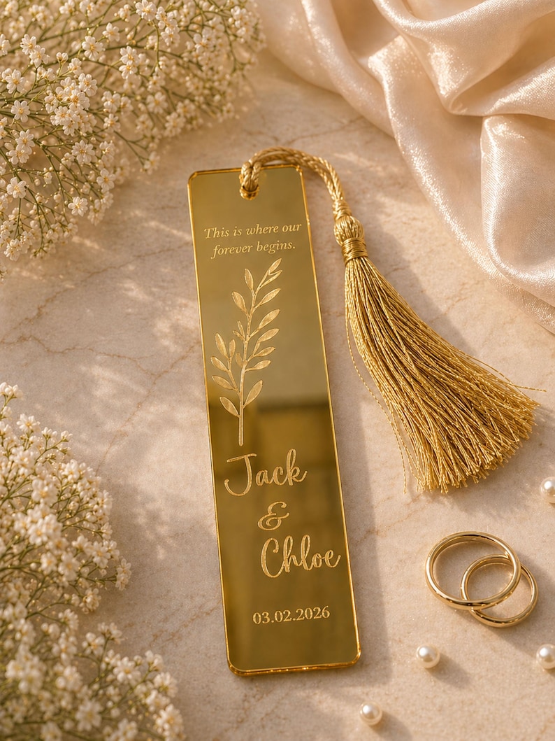 May include: A gold-colored bookmark with a tassel, featuring the engraved text "This is where our forever begins." and the names "Jack & Chloe" with the date 03.02.2026. Two gold rings and pearls are also visible.
