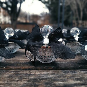 May include: A row of small, clear glass containers with a diamond-shaped top. Each container is decorated with a black ribbon and a small tag. The tag on the last container reads "HUSNUYE & M. ALI Nikah Hatırası".