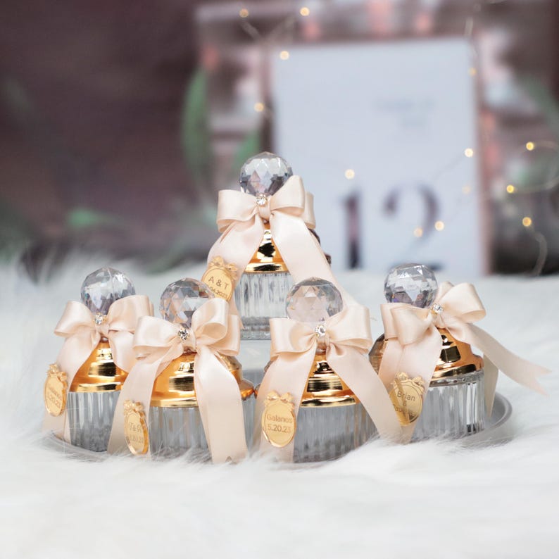 May include: Five clear glass jars with gold lids and a crystal-like knob on top. Each jar has a beige satin ribbon tied around the knob and a small gold tag with text on it. The jars are arranged in a pyramid shape on a white fluffy surface.