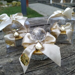 May include: Three clear glass boxes with gold lids and a crystal-like topper. Each box has a white satin ribbon and bow. The box in the center has a gold tag with the text "Our Wedding 10.02.2022".