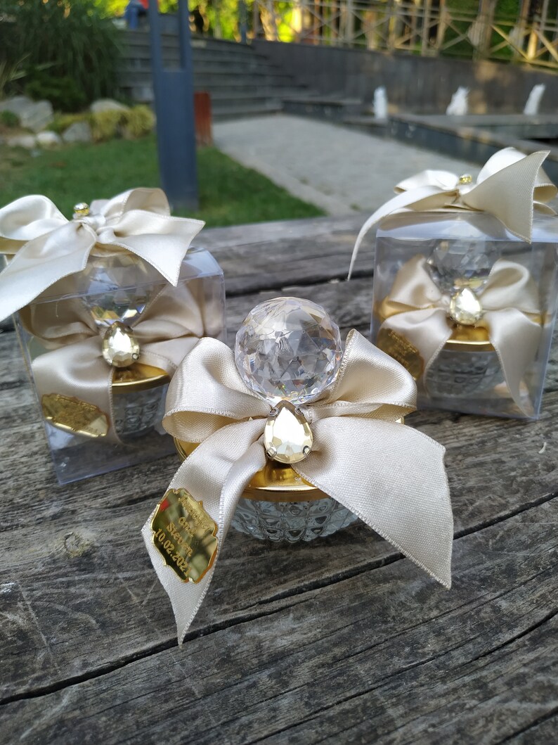 May include: Three clear glass boxes with gold lids and a crystal-like topper. Each box has a white satin ribbon and bow. The box in the center has a gold tag with the text "Our Wedding 10.02.2022".