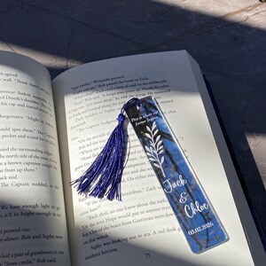 May include: A personalized bookmark with a blue and white design, featuring the names "Jack & Chloe" and the date 03.02.2026. The bookmark has a blue tassel and is placed in an open book.