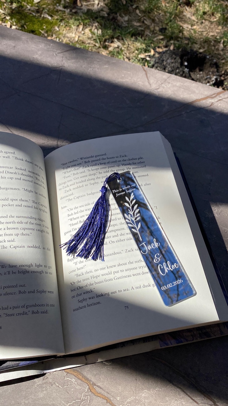May include: A personalized bookmark with a blue and white design, featuring the names "Jack & Chloe" and the date 03.02.2026. The bookmark has a blue tassel and is placed in an open book.