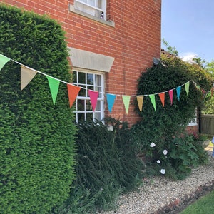 May include: A brick house with a green hedge and a string of colourful triangular flags hanging across the front. A young girl in a blue and white dress walks on a gravel path in the yard.