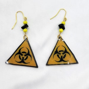 Puede incluir: Un par de pendientes de oro con cuentas amarillas y negras. Los pendientes tienen un colgante triangular con un símbolo de peligro biológico negro sobre un fondo amarillo.
