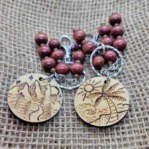 Puede incluir: Dos llaveros de madera con un diseño de montaña y una palmera. Los llaveros están unidos a una cadena de cuentas de madera marrón con anillos de llaves plateados.