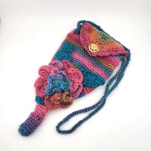 Op de afbeelding: Een handgemaakte gehaakte tas in levendige regenboogkleuren, met een grote, getextureerde bloemversiering in roze, blauwe en bruine tinten. De tas heeft een klepsluiting met een gouden knoop en een gevlochten band. De tas is ongeveer 20 cm hoog.