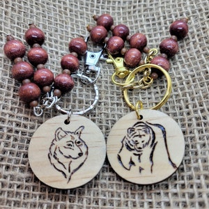 May include: Two wooden keychains with a wolf and a bear design. The keychains are attached to a brown wooden bead chain with a silver and gold key ring.