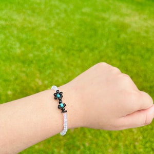 May include: A beaded bracelet featuring a floral design. The bracelet has a clear, beaded band with black and turquoise flower accents. The bracelet is worn on a wrist against a background of green grass.