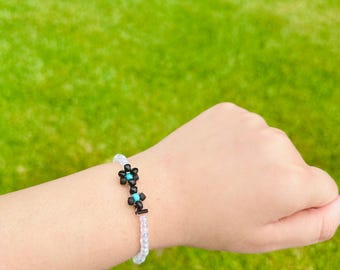 May include: A beaded bracelet featuring a floral design. The bracelet has a clear, beaded band with black and turquoise flower accents. The bracelet is worn on a wrist against a background of green grass.
