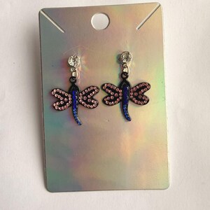 May include: A pair of dragonfly earrings featuring black metal frames, blue crystal bodies, and pink crystal wings. Each earring hangs from a clear round stud and is displayed on a rectangular, iridescent silver-toned cardboard backing card.